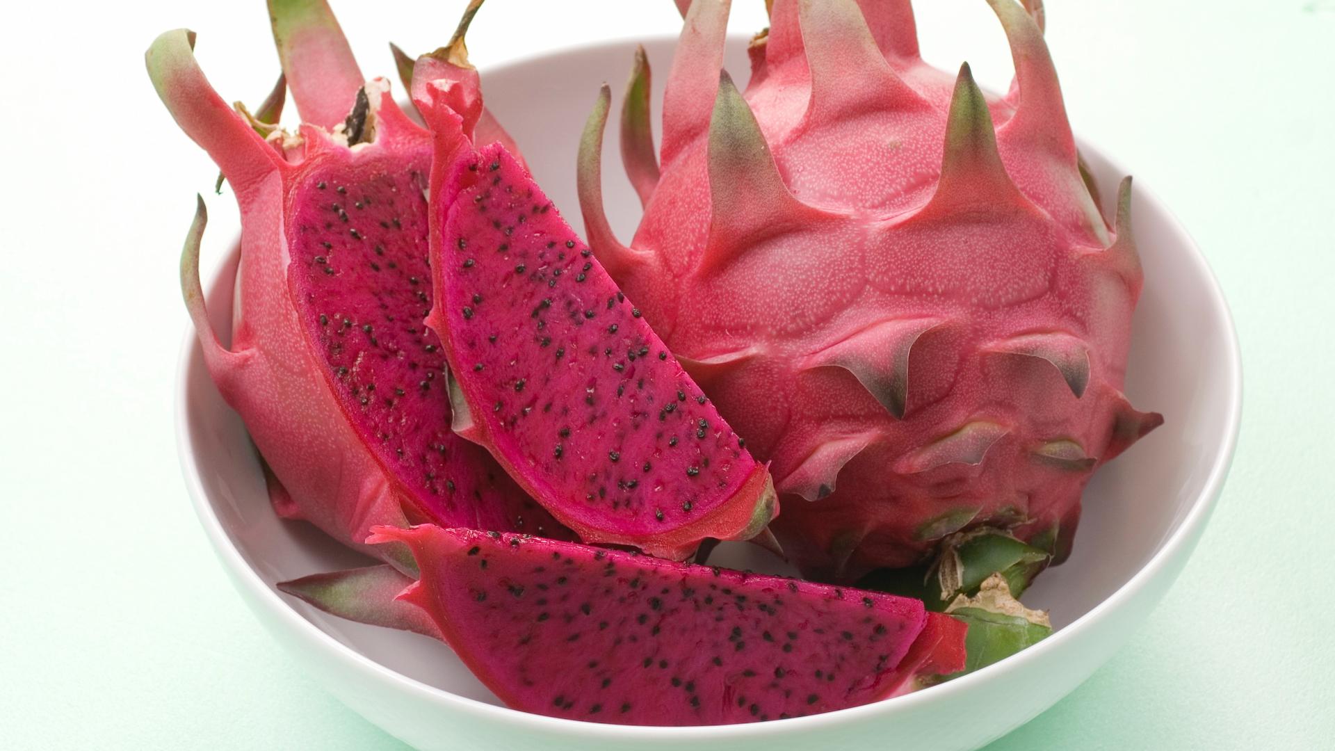 The image shows a white bowl containing several pieces of dragon fruit. Some of the fruit is whole, while other pieces have been cut to reveal the bright pink flesh and black seeds.
