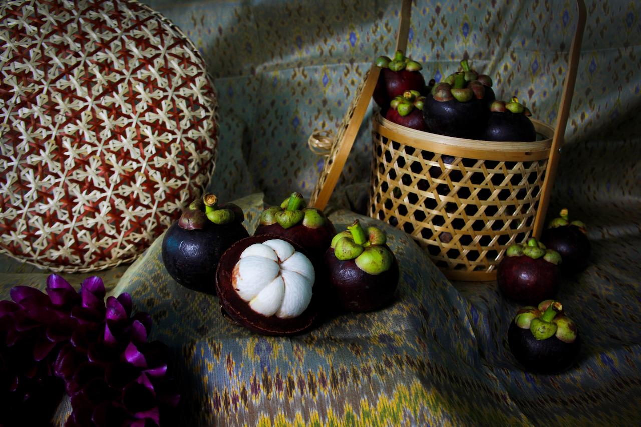 The image features a still life arrangement of mangosteens, some whole and some cut open to reveal the white fruit, along with a woven basket and a patterned background.  A decorative woven object and some purple flowers are also present.